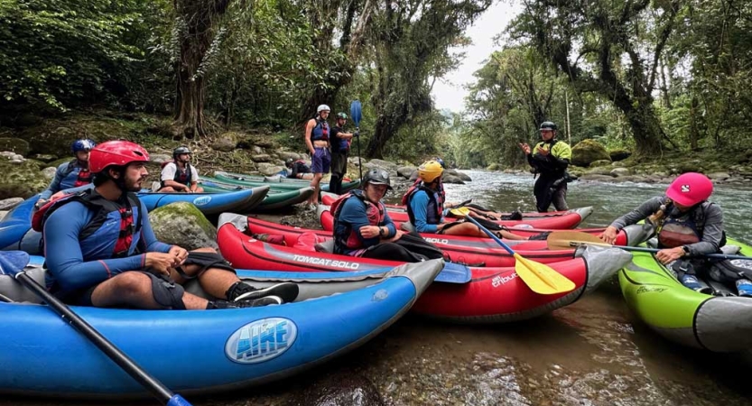 A group of people wearing safety get sit in small watercraft beached beside a river